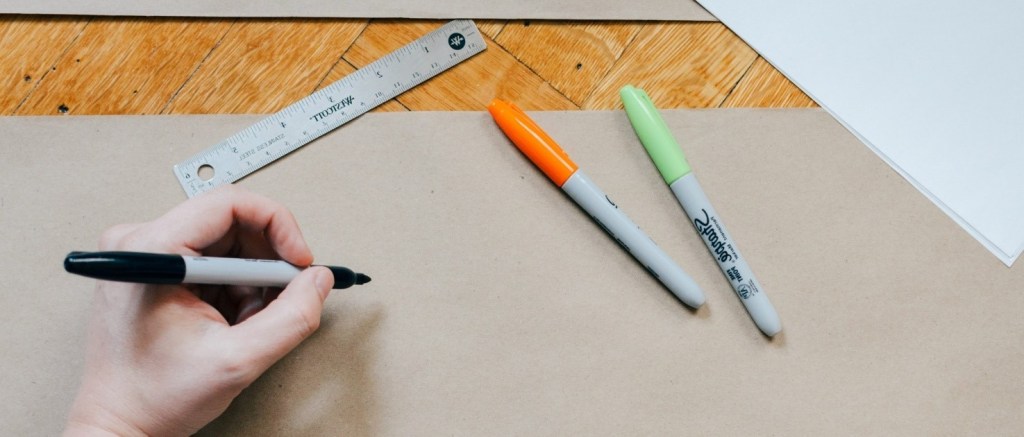 Una mano sostiene un lapiz en actitud de dibijar sobre un gran papel en blanco y sobre la mesa hay otras herramientas como lápices de colores, regla de dibujo, etc