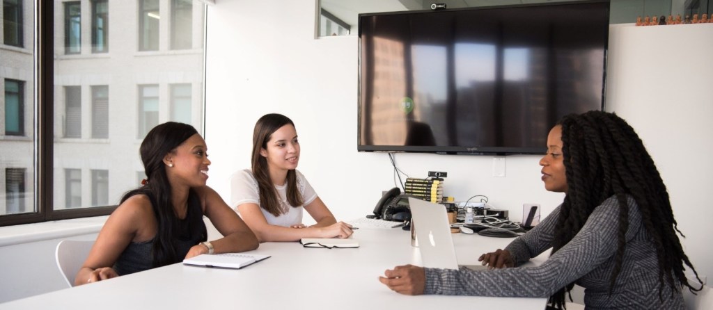 Dos mujeres reunidas en torno a una mesa de trabajo en un ambiente de oficina, escuchan las recomendaciones de una tercera.